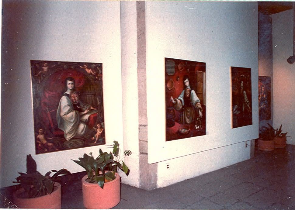 Sala de exposición con tres cuadros de la serie colgados sobre muro blanco y macetas en el suelo
